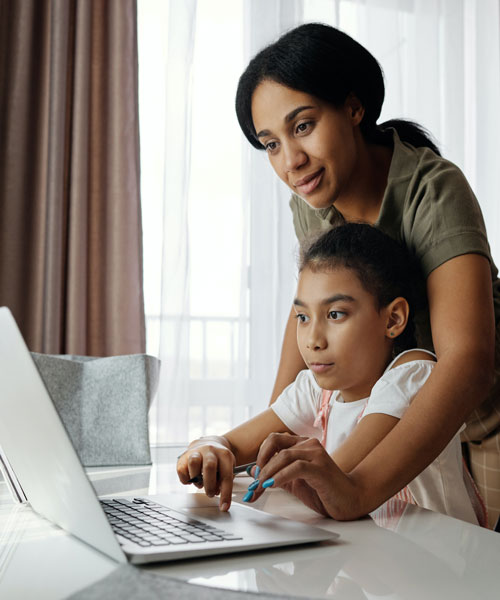 Older woman leaned over a young girl, helping her with homework on the laptop.